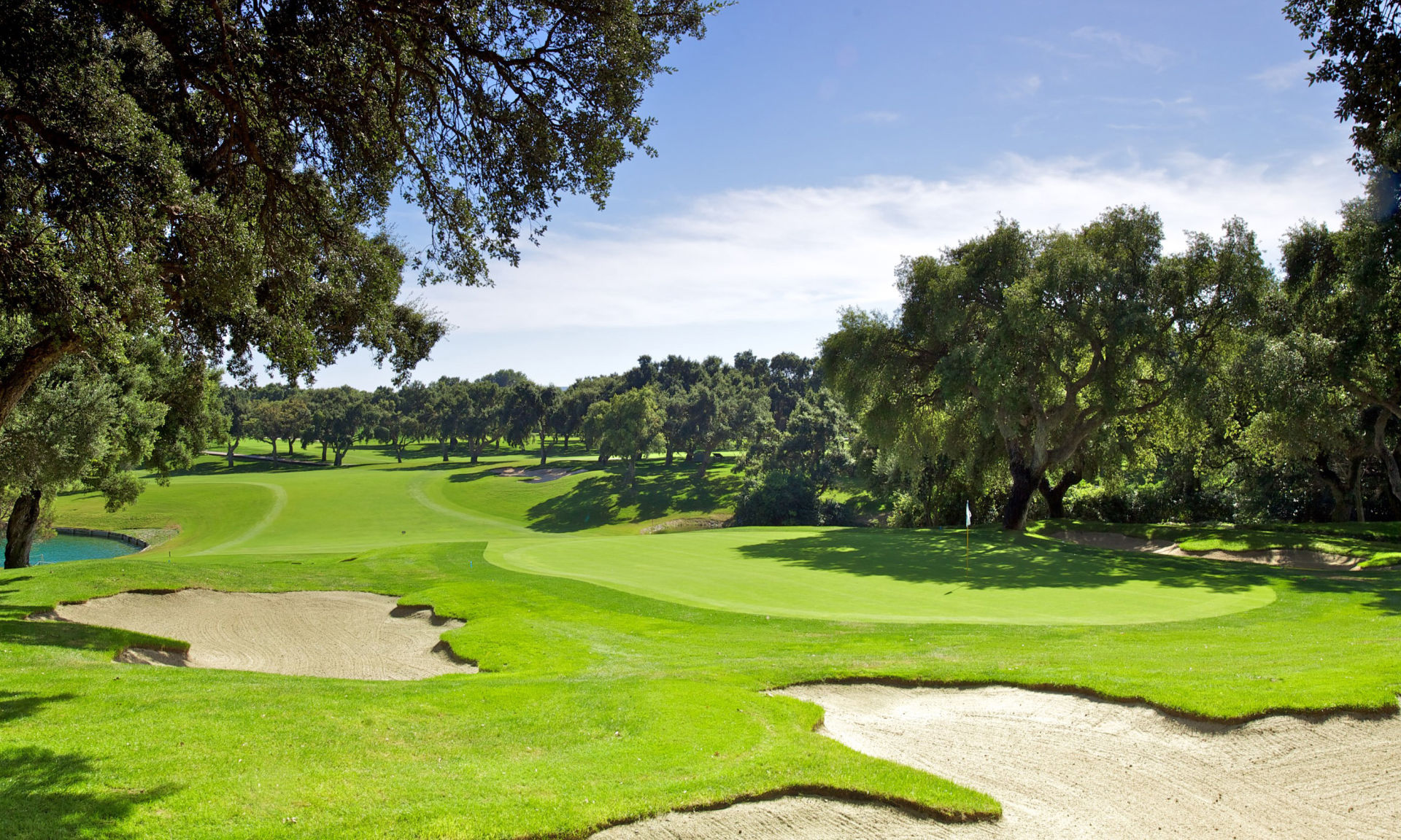 Vibrant view of Valderrama Golf Club showcasing lush green fairways, strategic sand bunkers, and a tranquil water hazard framed by mature oak trees under a clear sky.