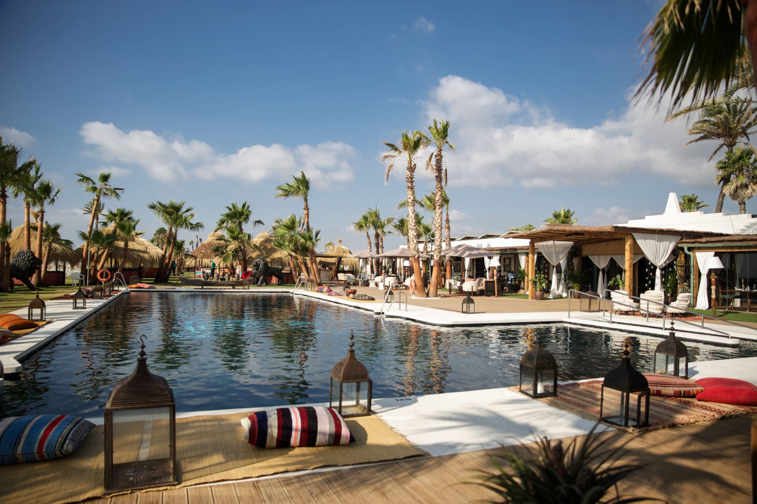 Luxurious beach club in Sotogrande featuring a serene pool surrounded by palm trees, thatched cabanas, and cozy seating with colorful cushions under a clear blue sky.
