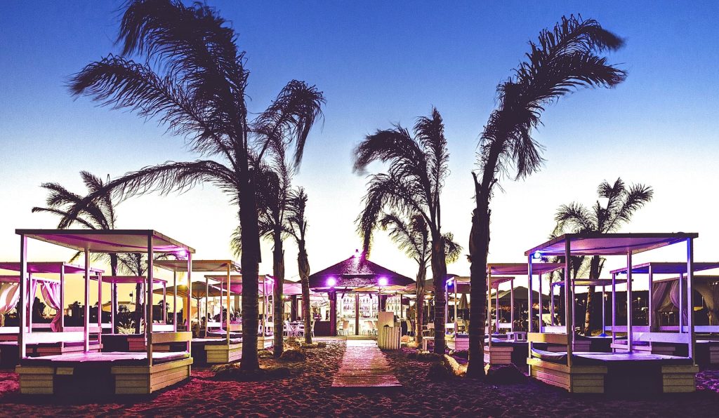 Evening view of a luxurious beach club in Sotogrande with modern cabanas, palm trees, and ambient purple lighting under a clear twilight sky.