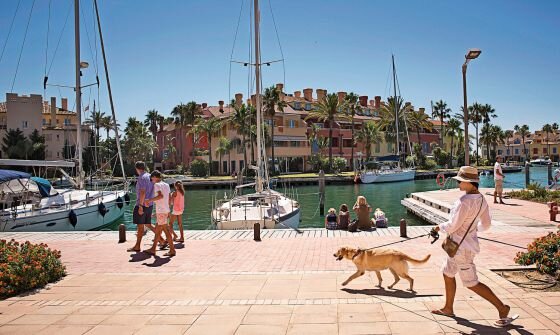 A scenic view of Sotogrande Marina with luxury yachts, waterfront apartments, and people enjoying a sunny day. A man in a white outfit walks his dog along the promenade.
